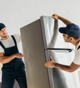 multicultural movers in uniform moving fridge in apartment